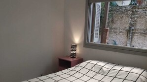 Een slaapkamer met een bed, nachtkastje, lamp en raam met uitzicht op een bakstenen muur en groen buiten.