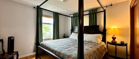 A four-poster bed with a floral bedspread, a bedside table with a lamp, and a chair.