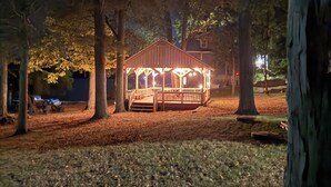 Ein Pavillon mit Holzkonstruktion und Dach, umgeben von Bäumen und einer Rasenfläche.