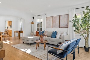 Een moderne woonkamer met een bank, salontafel en stoelen. Er is een eethoek met een tafel en stoelen. Een groot raam zorgt voor natuurlijk licht.