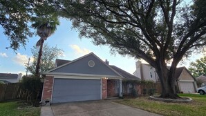 Garagem residencial com fachada de tijolos, uma árvore grande na frente e céu claro.