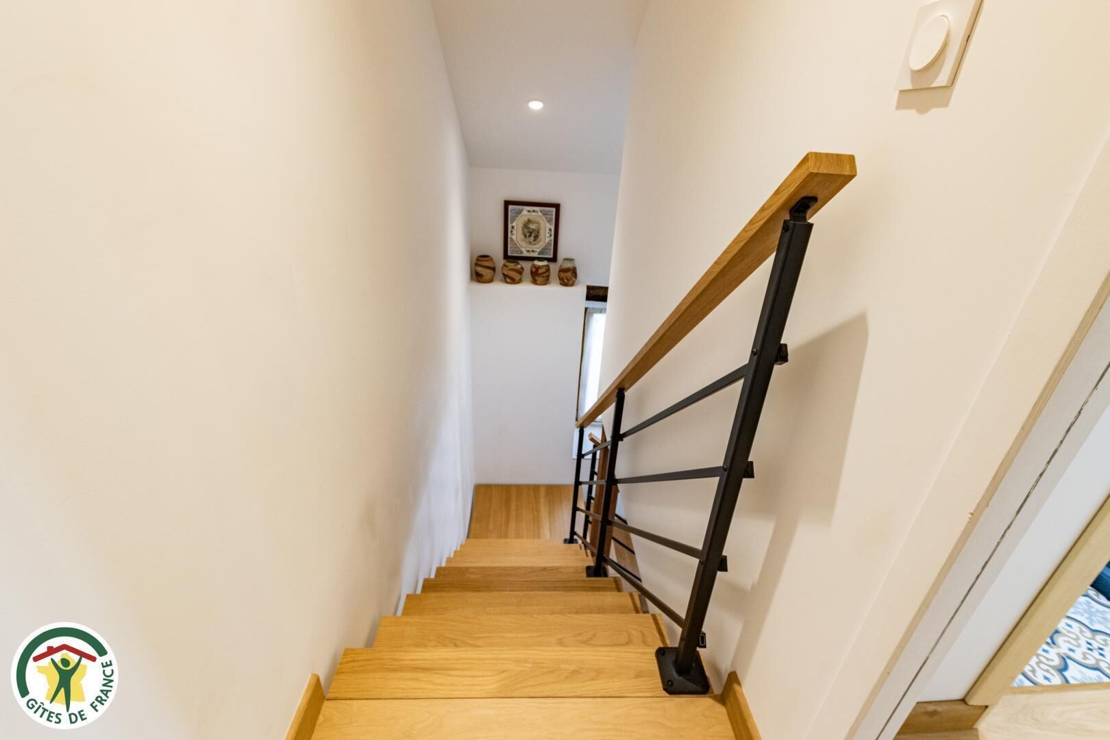 Un escalier en bois avec une rampe noire, menant à un palier orné d’un tableau encadré et d’objets décoratifs.