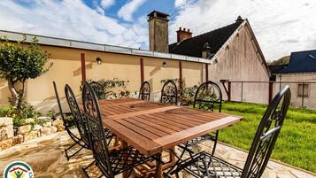 Patio esterno con tavolo di legno e sedie in metallo, circondato da pavimento in pietra e un'area verde.