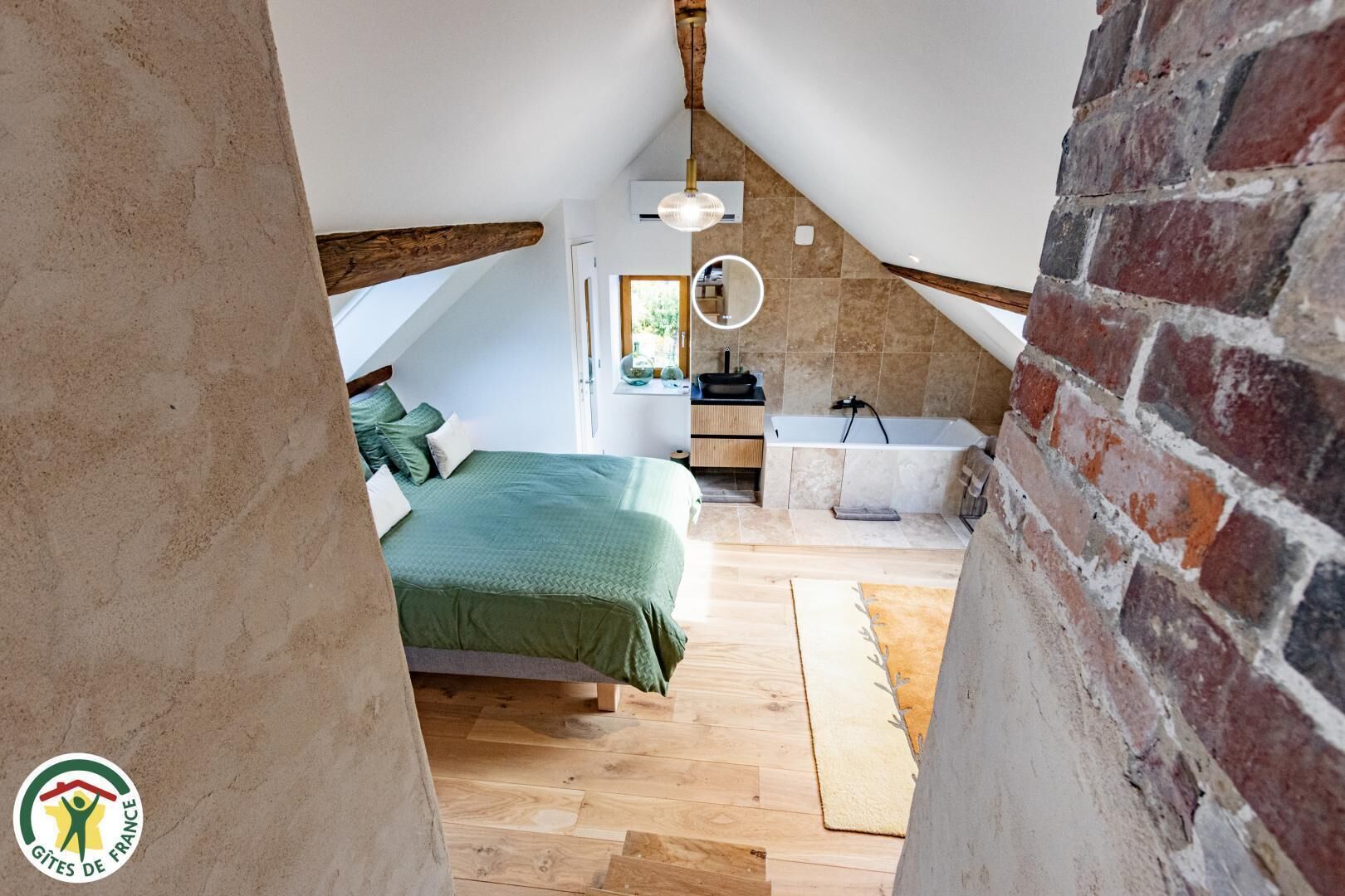 Una camera da letto con soffitto spiovente, un letto con biancheria verde, un comodino, uno specchio e un bagno con vasca.