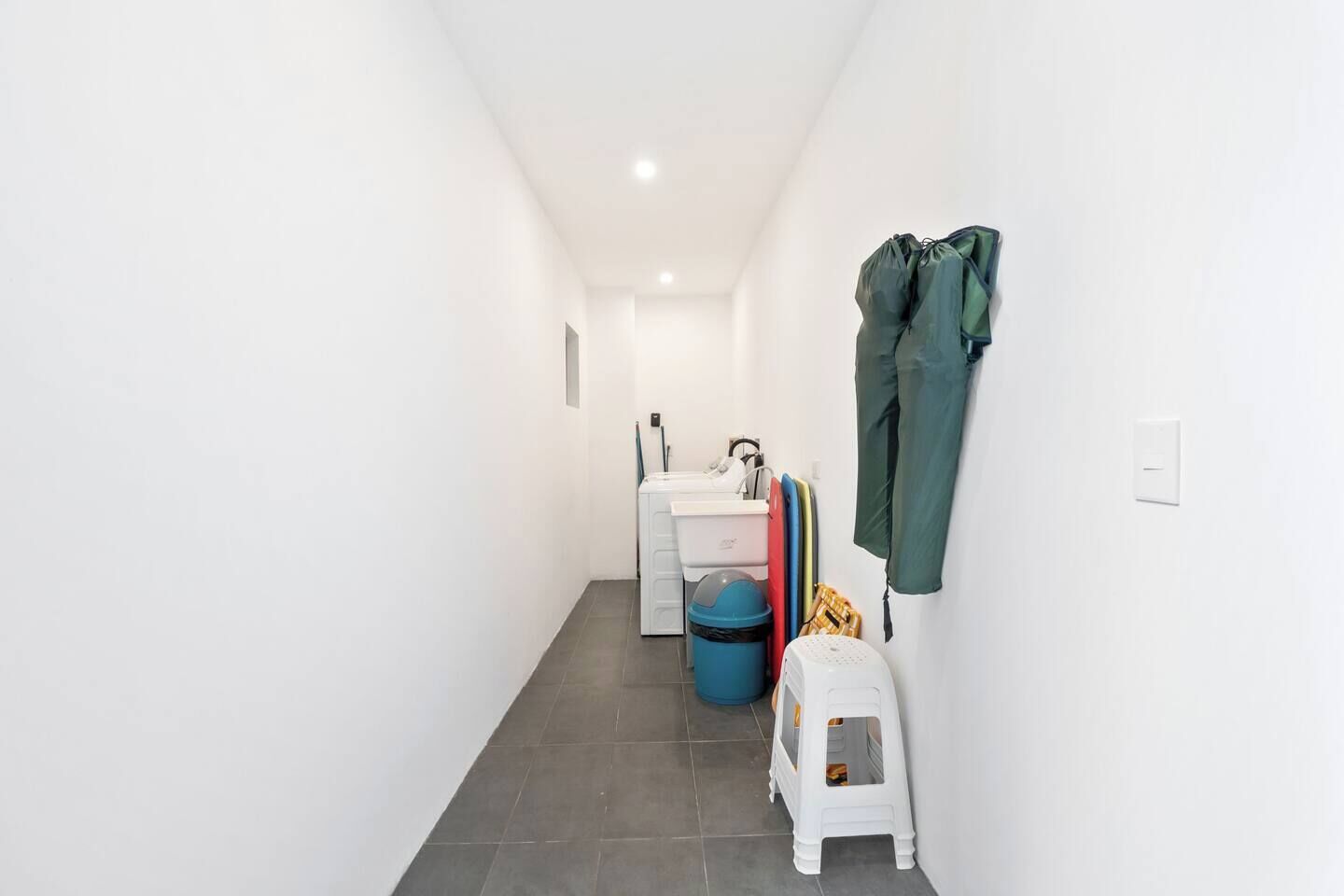 A narrow hallway with a white sink, a blue trash bin, and a small white stool.