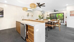 Eine moderne Küche mit einer zentralen Kochinsel, Holzschränken und einem eingebauten Geschirrspüler. An der Decke befinden sich Ventilatoren und Pendelleuchten über der Insel.