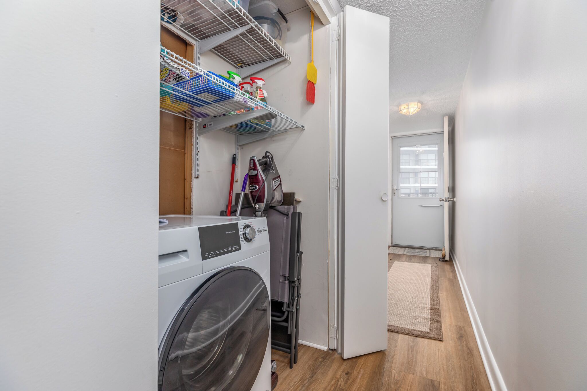 A laundry room with a front-loading washer and dryer, shelves with cleaning supplies, and a door leading to another room.