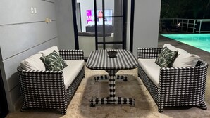 Um espaço de pátio com mesa e dois sofás xadrez, piso ladrilhado e piscina ao fundo.