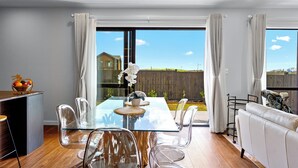 Una sala de estar moderna con una mesa de comedor de vidrio, sillas transparentes, un sofá y vista al jardín trasero a través de grandes ventanales.