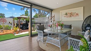 Un patio exterior con una puerta de vidrio que da a un jardín, una mesa redonda con sillas y un estante montado en la pared con adornos.