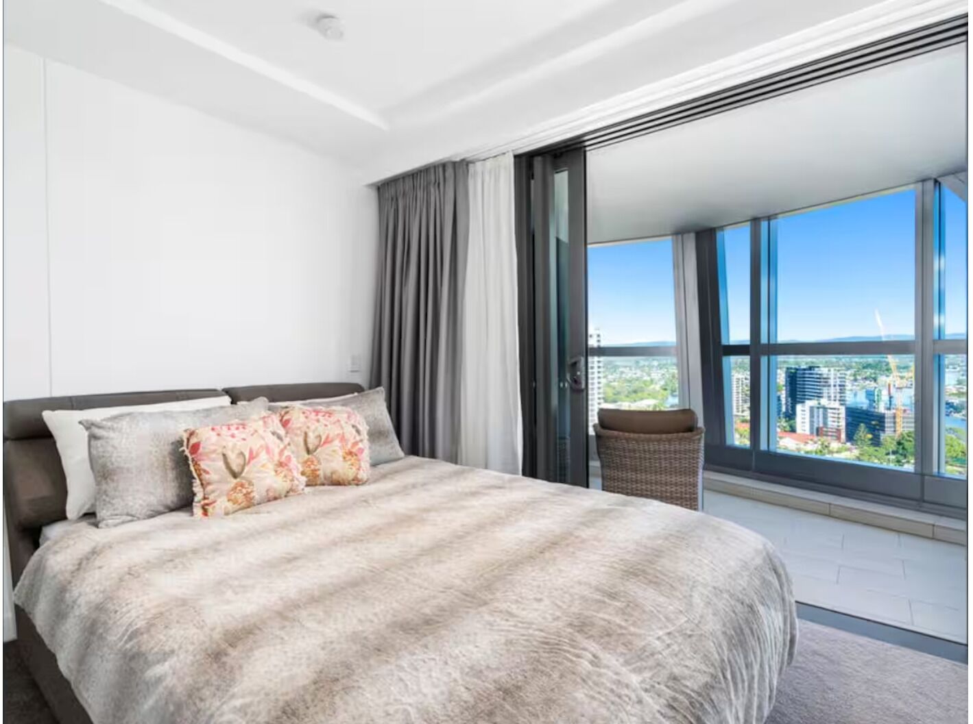 A bedroom with a large bed, a view of the city through floor-to-ceiling windows, and a balcony with a wicker chair.
