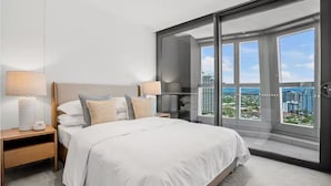 Une chambre moderne avec un grand lit, deux tables de chevet et une vue sur la ville grâce à de grandes fenêtres du sol au plafond.