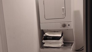 Una lavadora blanca y compacta, fijada a la pared, con un panel de control y un pequeño estante debajo para ropa doblada.