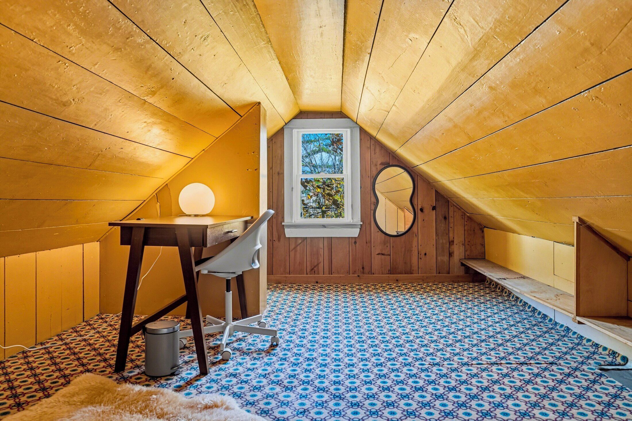 Una piccola stanza mansardata con soffitto spiovente, una scrivania con una sedia, una finestra e una chitarra appoggiata al muro.