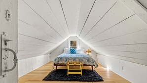 A narrow attic bedroom with a sloped ceiling, a bed with a blue and white patterned bedspread, a small wooden stool, and a black furry rug.