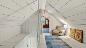 A narrow attic room with white wooden walls and a sloped ceiling. A beige sofa and a small table with a lamp are positioned against the wall. A blue rug lies on the floor, and two decorative wall panels are mounted on the wall.
