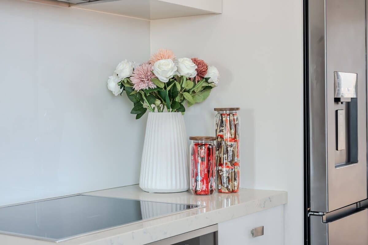 A kitchen counter with a white vase containing flowers, two glass jars with red contents, and a stainless steel refrigerator.