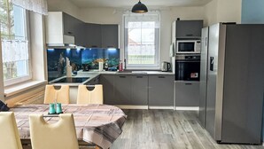A modern kitchen with grey cabinets, a stainless steel refrigerator, a microwave, and a sink. There is a dining table with chairs and a window with curtains.