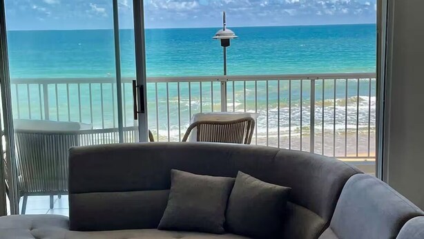 A modern living room with a large sectional sofa, a small table, and a patterned rug. The room has a large sliding glass door leading to a balcony with a view of the ocean and a distant island. - Beachfront 3-Bedroom Apartment with Breathtaking Views in Luquillo (Luquillo)