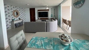 A modern living room with a grey sofa, a dining table with a teal table runner, and a bowl of decorative stones. A large wall clock and a mirror are visible. - Beachfront 3-Bedroom Apartment with Breathtaking Views in Luquillo (Luquillo)