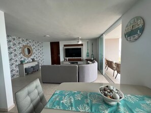 A modern living room with a grey sofa, a dining table with a teal table runner, and a bowl of decorative stones. A large wall clock and a mirror are visible. - Beachfront 3-Bedroom Apartment with Breathtaking Views in Luquillo (Luquillo)