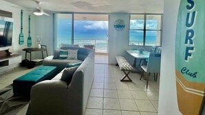 A living room with a view of the ocean, a surfboard decoration, and a dining area. - Beachfront 3-Bedroom Apartment with Breathtaking Views in Luquillo (Luquillo)
