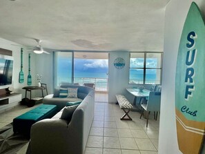 A living room with a view of the ocean, a surfboard decoration, and a dining area. - Beachfront 3-Bedroom Apartment with Breathtaking Views in Luquillo (Luquillo)