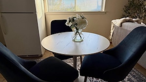 Un comedor con una mesa redonda, dos sillas, un jarrón con flores, un reloj en la pared, una ventana con persianas y una planta.