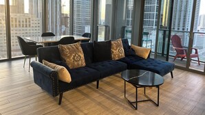 Een moderne woonkamer met een donkerblauwe hoekbank, een ronde salontafel, en een eethoek met een tafel en stoelen. Grote ramen bieden uitzicht op hoge gebouwen.