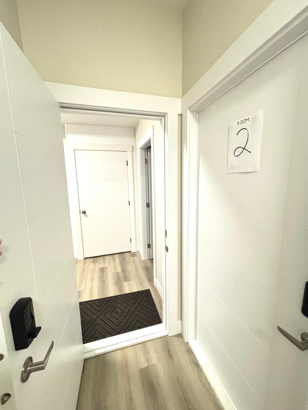 Un couloir avec une porte sur laquelle est inscrit « SALLE 2 », un paillasson noir et un plancher en bois de couleur claire.