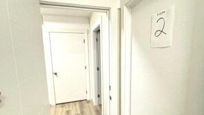 Un corridoio con una porta con la scritta 'ROOM 2', un tappetino nero e un pavimento in legno chiaro.