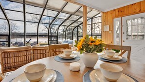 A dining area with a table set for four, surrounded by wicker chairs, a vase with sunflowers, and a view of a snowy landscape through large windows. - Lake Views | Tub Sauna Kayak Bike Fire Pit EV (Albrightsville)