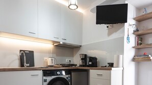 Eine kompakte Küche mit Waschmaschine, Kaffeemaschine und einem flachbildfernseher an der Wand.
