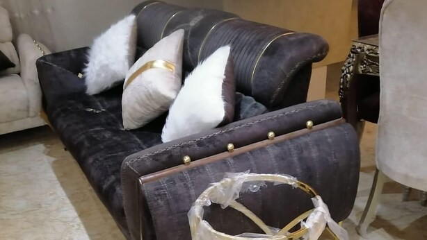 Een donkerbruine bank met witte en gouden kussens, een marmeren vloer en een stoel met een witte hoes.