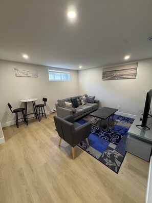 A modern living room with a grey sofa, a coffee table, and a TV stand. There is a dining area with a table and chairs, and artwork on the walls.