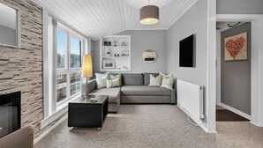 A modern living room with a grey sofa, a black coffee table, and a floor lamp. There is a fireplace on the left, a white wall with shelves, and a large window. - Coastal Charm: Modern Retreat by Seafront (Largs)