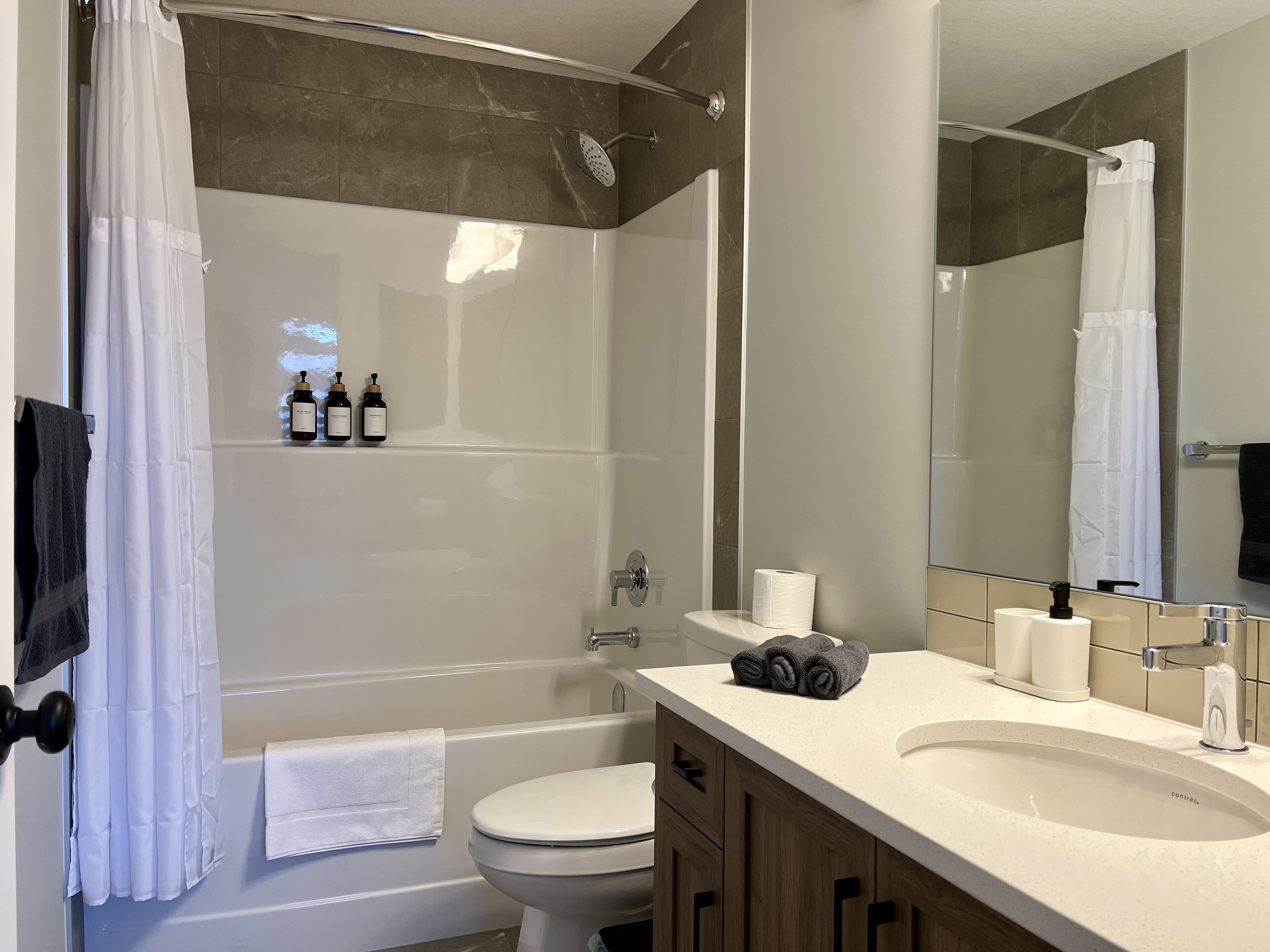 Une salle de bain moderne avec une baignoire, des toilettes et un lavabo. On y trouve des bouteilles de produits de toilette fixées au mur, ainsi qu’un porte-serviettes.