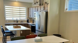 A modern kitchen with a central island, stainless steel appliances, and a dining area with a white table and colorful chairs. - Modern 3BR | Bonus Room | Office | A/C, Smart TVs, Parking (Calgary)