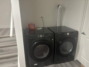Two black Samsung washing machines in a laundry room with a white door and a wooden floor. - Peniel Place - Your Peaceful Home Away From Home (Airdrie)