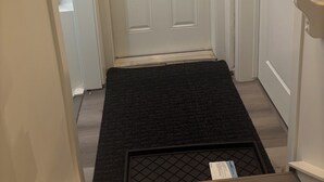 Eine weiße Tür mit Zahlenschloss, eine schwarze Fußmatte im Eingangsbereich und ein kleines Regal zur Rechten.