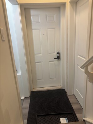 A white door with a keypad lock, a black mat at the entrance, and a small shelf to the right. - Peniel Place - Your Peaceful Home Away From Home (Airdrie)