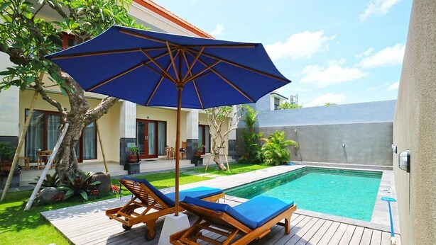 A poolside area with a wooden deck, two lounge chairs, a blue umbrella, and a small table. - Lotus Canggu (Tibubeneng)