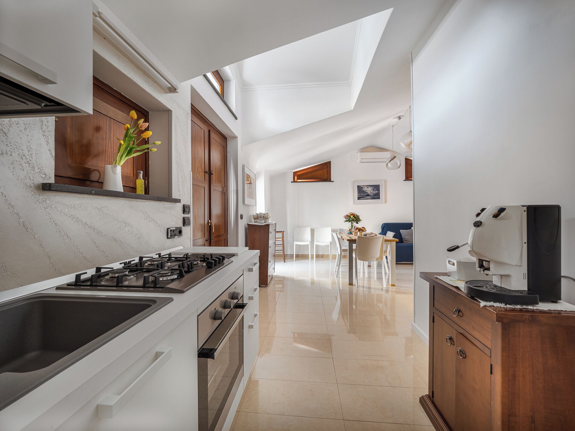 A modern kitchen with a gas stove, oven, and a coffee machine. There is a dining area with a table and chairs, and a blue sofa.
