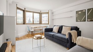 Ein modernes Wohnzimmer mit einem dunkelblauen Sofa, einem weißen Teppich und einem Holztisch. Es gibt einen Esstisch mit Stühlen und ein großes Fenster mit Jalousien.
