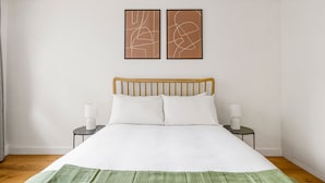 Ein ordentlich bezogenes Bett mit weißer Bettwäsche und einer grünen Tagesdecke, zwei Nachttischlampen und abstrakte Wandbilder.