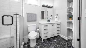 A modern bathroom with a white vanity, a toilet, a shower with a glass door, and a black and white patterned floor. - Uptown Vibe, Downtown Location - Sleeps 6 (Indianapolis)
