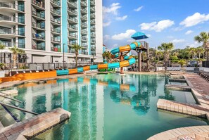 A hotel pool with a water slide, a multi-story building with balconies, and palm trees. - Last Chance Summer, Oceanfront Pools and Beach (Myrtle Beach)
