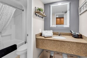 A bathroom with a white toilet, a sink with a granite countertop, a mirror above the sink, and a shower area with a white shower curtain. - Last Chance Summer, Oceanfront Pools and Beach (Myrtle Beach)