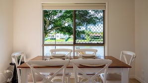 Ein Essbereich mit Holztisch und weißen Stühlen, ein Fenster mit Blick auf ein Haus und Bäume sowie eine Steckdose an der Wand.