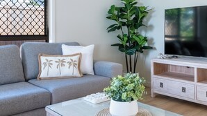 Ein modernes Wohnzimmer mit grauem Sofa, weißem Couchtisch, einer großen Topfpflanze, einem Fernseher auf einem Holzständer und einem Fenster mit einem Metallgitter.